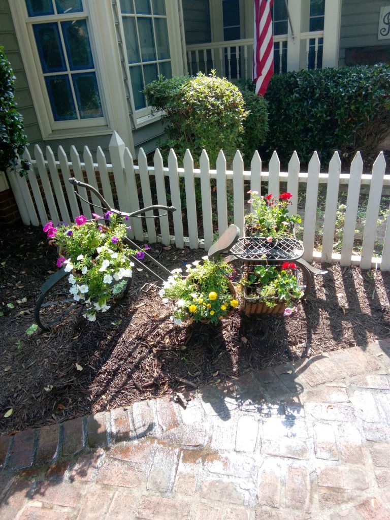 flower garden in front of a house with a white picket fence and a bicycle with flowers